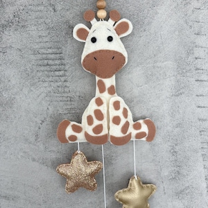 Könnte beinhalten: Ein Baby-Mobile mit einer Giraffe aus Filz mit braunen Flecken und Akzenten, das an einer Schnur mit Holzperlen hängt. Drei goldene Stern-Ornamente hängen darunter. Die Giraffe hat einen weißen Körper und braune Details.