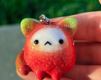 May include: A handmade red and white charm shaped like a cartoon animal with black eyes and a small black mouth. The charm has green accents on the ears and a silver metal loop for attaching to a keychain or necklace.