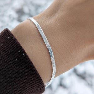 May include: A silver herringbone bracelet is shown on a wrist. The bracelet is made of small, flat, interlocking links, creating a smooth, flexible band. The bracelet is a bright, shiny silver color. The background is blurred, with a snowy appearance.