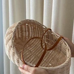 Puede incluir: Una cesta de mimbre beige claro con un asa de cuero marrón. La cesta tiene forma ovalada y presenta un patrón tejido.