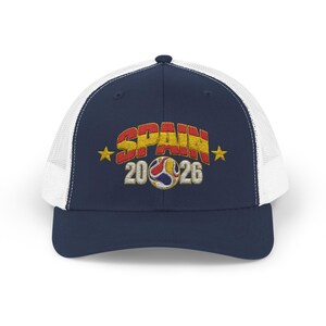 May include: A navy blue trucker hat with a white mesh back. The front features embroidered text that reads SPAIN in red and yellow, with the number 2026 below it. A soccer ball graphic sits between the numbers, flanked by two yellow stars.