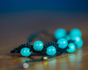 Pode incluir: Um bracelete de contas turquesa com cordão preto. As contas são redondas e têm uma superfície ligeiramente irregular.