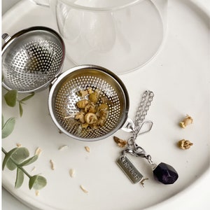 May include: A stainless steel tea infuser ball with a chain and a charm that says "enough" and a purple amethyst stone. The infuser ball is filled with dried chamomile flowers.