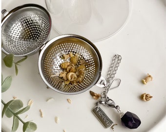 May include: A stainless steel tea infuser ball with a chain and a charm that says "enough" and a purple amethyst stone. The infuser ball is filled with dried chamomile flowers.