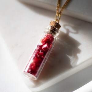 May include: A gold chain necklace with a small glass vial pendant filled with red coral chips. The vial has a cork stopper.