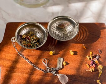 May include: A silver tea infuser with a chain and a pink crystal charm. The infuser is open and filled with dried herbs. The infuser is on a wooden surface with dried herbs scattered around it. The tag on the chain reads "PROTECTED".