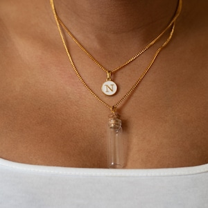 May include: A gold chain necklace with two pendants. The first pendant is a round white disc with the letter "N" in gold. The second pendant is a small, clear glass bottle with a cork stopper.