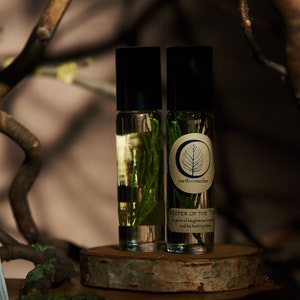 May include: Two small glass bottles with black caps and labels. The bottles contain a clear liquid and green plant matter. The label on the right bottle reads "earth.remedies" and "Keeper of the Trees to ground heightened emotions and be held by the trees".