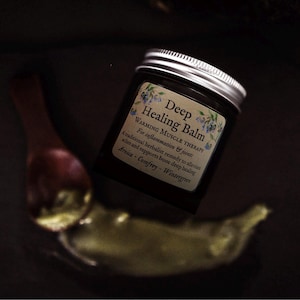 Könnte beinhalten: Ein dunkles Glasgefäß mit silbernem Schraubdeckel und einem Etikett mit der Aufschrift Deep Healing Balm, Warming Muscle Therapy, for inflammation and joints, a traditional herbalist remedy to alleviate aches and supports bone deep healing, Arnica, Comfrey, Wintergreen, begleitet von einem kleinen Holzlöffel mit einem Klecks grünlichem Balsam.