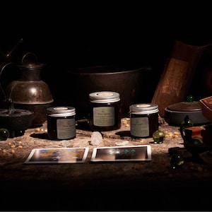 May include: Three amber glass jars with silver lids and white labels, each labeled Three Lights, arranged alongside two rectangular cards, clear crystals, and small green spheres on a dark, textured surface with various metal vessels.