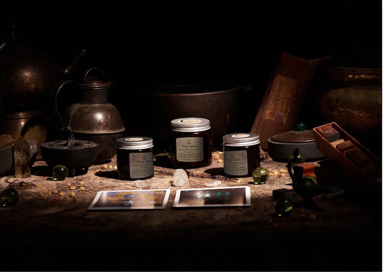 May include: Three amber glass jars with silver lids and white labels, each labeled Three Lights, arranged alongside two rectangular cards, clear crystals, and small green spheres on a dark, textured surface with various metal vessels.