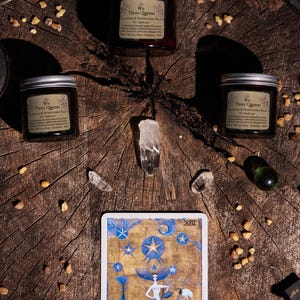 May include: Three amber glass jars with silver lids labeled We Three Queens Cleansing & Moisturizing Balm, arranged around clear quartz crystals and a tarot card titled The Star, featuring a blue and gold illustration of a figure under stars.