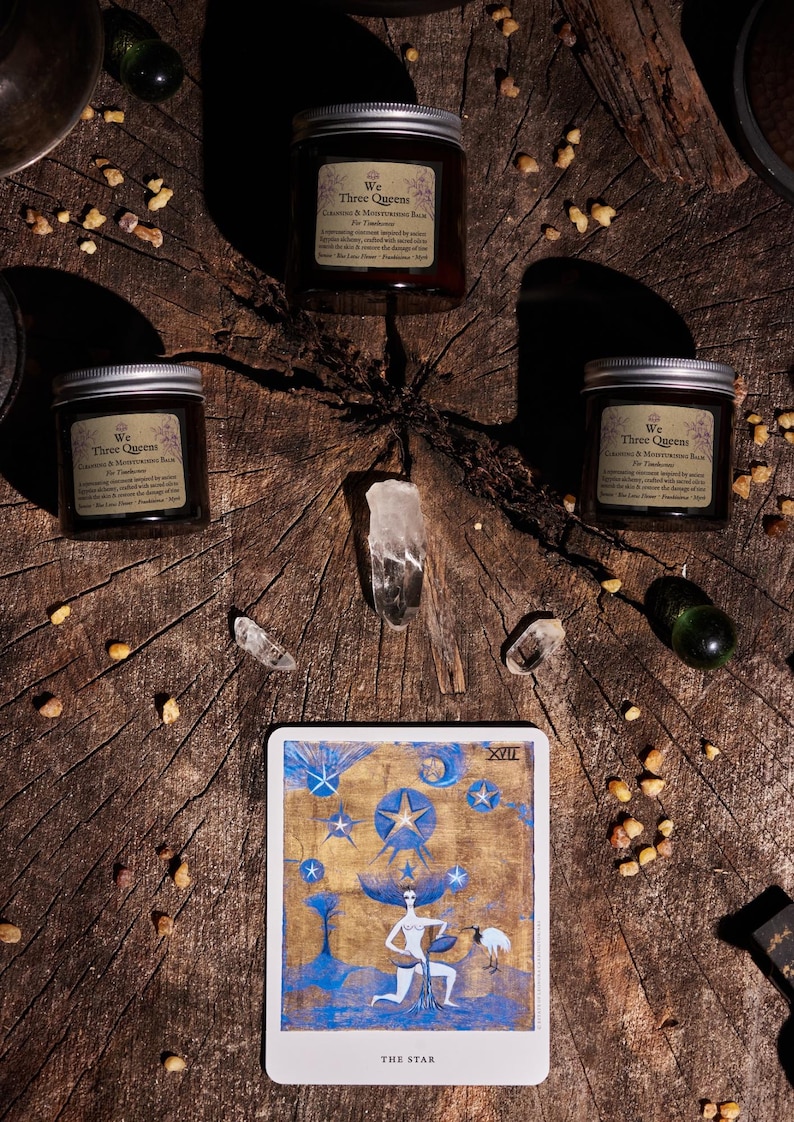 May include: Three amber glass jars with silver lids labeled We Three Queens Cleansing & Moisturizing Balm, arranged around clear quartz crystals and a tarot card titled The Star, featuring a blue and gold illustration of a figure under stars.
