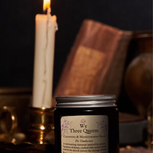 May include: A dark amber glass jar with a silver screw-top lid featuring a label that reads We Three Queens Cleansing & Moisturising Balm For Timelessness, A rejuvenating ointment inspired by ancient Egyptian alchemy, Jasmine, Blue Lotus Flower, Frankincense, Myrrh.