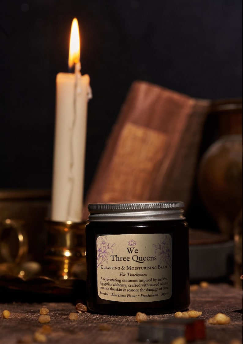 May include: A dark amber glass jar with a silver screw-top lid featuring a label that reads We Three Queens Cleansing & Moisturising Balm For Timelessness, A rejuvenating ointment inspired by ancient Egyptian alchemy, Jasmine, Blue Lotus Flower, Frankincense, Myrrh.