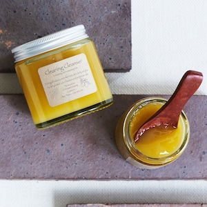 Op de afbeelding: Een glazen pot met gele gezichtsreiniger en een zilveren deksel. De pot is gelabeld met "Clearing Cleanser" en vermeldt ingrediënten zoals tea tree en citroen. De pot staat op een bruine tegelvloer.