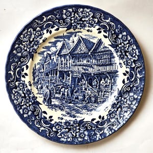Puede incluir: Un plato decorativo redondo, azul y blanco, con una detallada ilustración de una escena de ciudad histórica. El borde del plato está adornado con un patrón floral, creando una estética clásica y vintage.