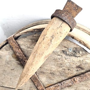 Puede incluir: Una herramienta de madera vintage con forma cónica y una banda y cuchilla de metal oxidado. La herramienta se encuentra sobre una superficie de madera desgastada con bandas de metal oxidadas, lo que sugiere una estética antigua o rústica.