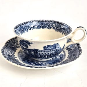 Könnte beinhalten: Ein Vintage-Tee-Set, bestehend aus einer Tasse und einer Untertasse. Die Tasse und die Untertasse sind weiß mit einem blauen Transferdruck-Design mit einer Landschaftsszene. Der Rand der Tasse und der Untertasse hat eine dekorative Bordüre. Die Tasse hat einen gebogenen Henkel.