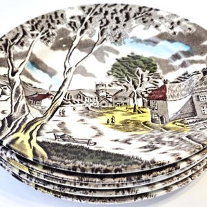 May include: A stack of vintage ceramic plates featuring a black and white pastoral scene with a church, trees, and houses. The plates have a cream-colored border and a yellow and blue sky.
