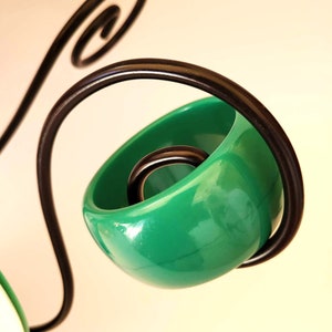 Peut inclure: Gros plan sur un objet vert brillant en forme de bol avec un anneau sombre à l'intérieur, maintenu par un cadre métallique noir incurvé. Le cadre a une volute décorative en haut. Le fond est de couleur blanc cassé.