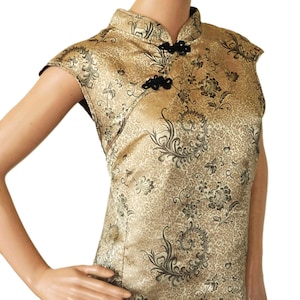 May include: A sleeveless gold dress featuring a black floral and scroll pattern. The garment has a high mandarin collar and a side closure secured with two black fabric knot buttons. The fabric has a subtle sheen and a textured, woven appearance.