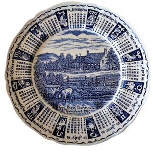 May include: A blue and white ceramic plate with a calendar design for each month of the year. The plate features a cottage scene with a blue and white colour scheme. The text "God Bless Our House Throughout 1985" is printed on the plate.