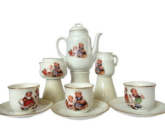 May include: A white porcelain tea set with a whimsical design featuring children playing. The set includes a teapot, creamer, sugar bowl, and four teacups and saucers.