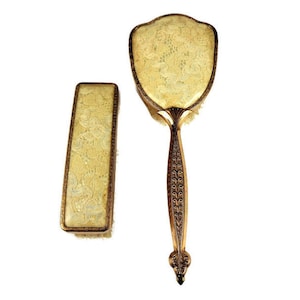 Könnte beinhalten: Ein Vintage-Handspiegel- und Bürstenset in Goldtönen mit floralem Muster. Der Spiegel hat einen gewellten Rand und die Bürste einen langen Stiel.