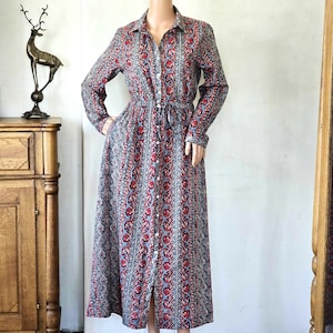 Puede incluir: Un vestido largo de manga larga con botones y estampado floral en rojo, azul y gris. El vestido tiene cuello camisero, cintura anudada y falda larga. El vestido se exhibe en un maniquí.