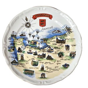 May include: A white ceramic decorative plate featuring a colourful illustrated map of the Normandie region in France. The design includes a compass rose, the text NORMANDIE and MANCHE, various landmarks, ships, and parachutes across the map surface.