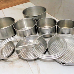 Könnte beinhalten: Ein Set silberfarbenes Kochgeschirr aus Metall, bestehend aus Töpfen, Pfannen und Deckeln. Das Kochgeschirr ist auf einer weißen gefliesten Oberfläche angeordnet.