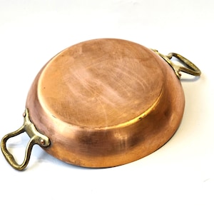 May include: A copper-colored, round pan with two brass handles. The pan has a shiny, reflective surface and a slightly curved shape. The handles are attached to the sides of the pan and are curved for easy gripping. The pan is likely used for cooking.