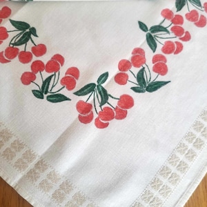 Può includere: Tovaglia bianca con un bordo di piccoli quadrati e un motivo ripetuto di ciliegie rosse e foglie verdi.