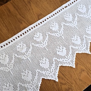May include: A white crochet valance featuring a repeating floral pattern of tulips. The fabric has a delicate, open-work mesh construction with a scalloped bottom edge and a straight top border with a series of small, evenly spaced circular cutouts.