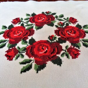 May include: A white fabric with a red rose wreath embroidered on it. The roses are in a circle with green leaves.