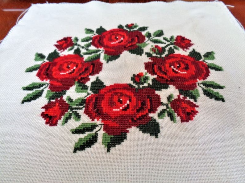 May include: A white fabric with a red rose wreath embroidered on it. The roses are in a circle with green leaves.