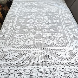 Puede incluir: Un mantel blanco, intrincadamente tejido a ganchillo, con un patrón floral y geométrico. El mantel rectangular muestra un diseño calado detallado. Los bordes presentan un borde decorativo.