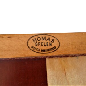 May include: Close-up of a wooden game board with a brown and white checkered pattern. The board has a label that reads "Homas Spelen" in a black oval.
