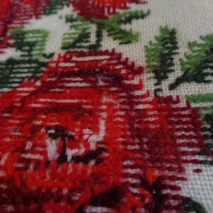 May include: Close-up of a red and green embroidered fabric with a floral design. The fabric is woven with a textured pattern and features a repeating motif of red roses and green leaves.