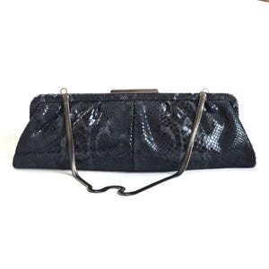 Pode incluir: Uma bolsa clutch preta com um padrão texturizado de pele de cobra. A bolsa retangular possui um fecho de estrutura metálica no topo e uma alça de corrente metálica fina e flexível presa a dois pequenos anéis nos cantos superiores da estrutura.