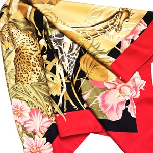 May include: A silk scarf with a vibrant animal and floral print. The design features a leopard, giraffe, and pink flowers against a black, gold, and red background. The scarf has a red border.