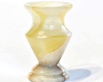 May include: A small, polished stone vase featuring a smooth, translucent surface with swirling patterns of cream, pale yellow, and light brown. The vessel has a wide, flared rim, a tapered neck, a rounded body, and a sturdy, circular base.