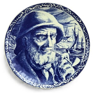May include: A blue and white decorative plate depicting an elderly man with a beard, wearing a hat and smoking a pipe. The background shows a coastal scene with boats and buildings. The plate has a decorative border.