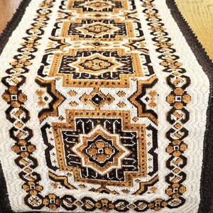 May include: A rectangular table runner featuring a repeating geometric pattern in shades of dark brown, golden yellow, and cream. The design includes central star-like motifs surrounded by intricate borders and finished with dark brown fringe edges.