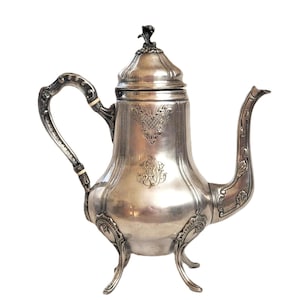Puede incluir: Una tetera de plata con un diseño ornamentado y tres patas. La tetera tiene una tapa con un pomo y un pico. La tetera está decorada con motivos florales grabados.