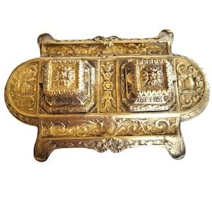 May include: A gold-toned metal inkwell with ornate detailing and two inkwells. The inkwell has a rectangular shape with a raised, decorative border. The inkwells are square and have a raised, decorative border. The inkwell is on a white background.