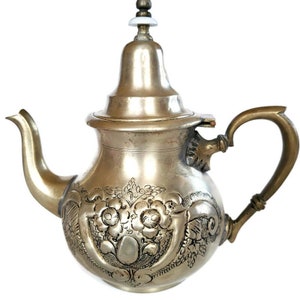 Puede incluir: Una tetera de plata con un diseño floral ornamentado y una tapa con bisagras. La tetera tiene un cuerpo redondo y un pico curvo. El asa es de latón.