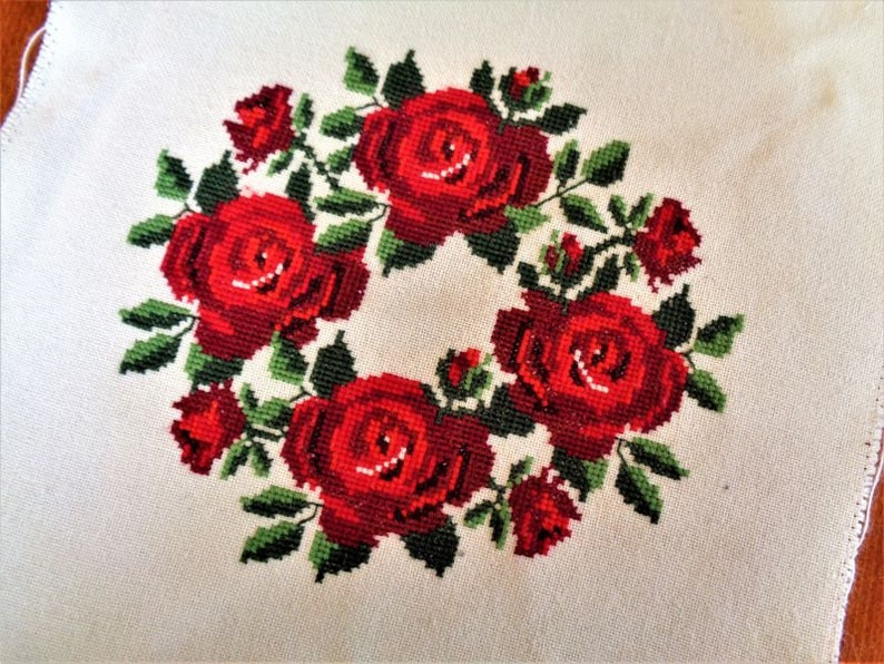 May include: A white fabric with a red embroidered wreath of roses. The roses are in a circular pattern with green leaves.