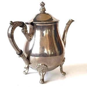 May include: A silver teapot with a decorative lid and ornate handle. The teapot has three legs and a bulbous body.